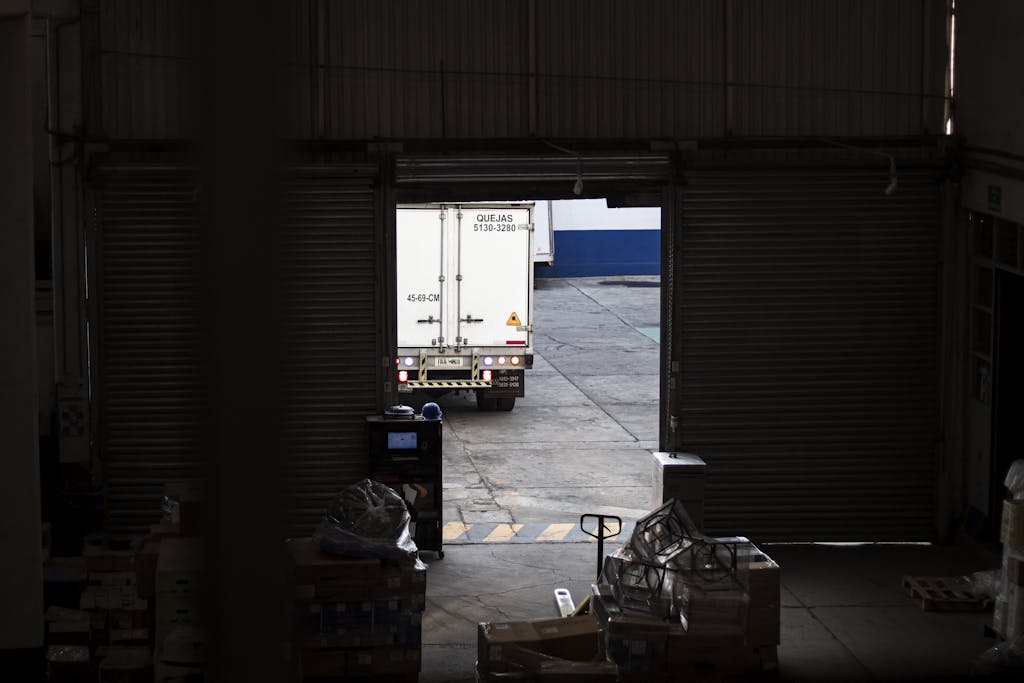 A white delivery truck exits a dimly lit warehouse loading dock, surrounded by packages and pallets.